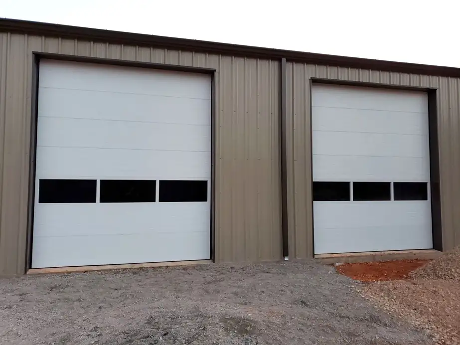 Two white commercial doors with black vision panels on a tan steel building, installed by Integrity Overhead Door