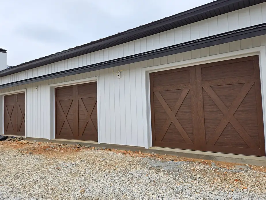 Three woodgrain X-brace carriage doors along a white board-and-batten building, installed by Integrity Overhead Door